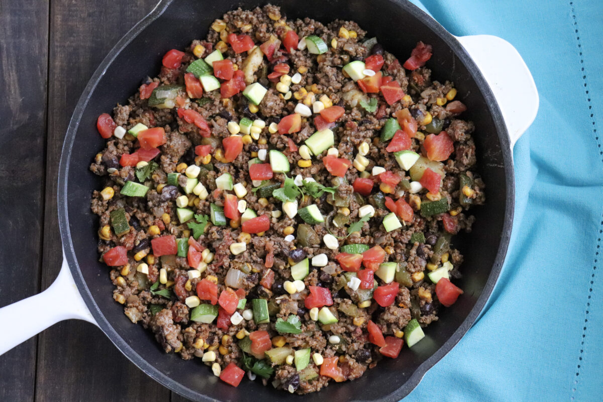 cast iron skillet with cooked hamburger meat and visible pieces of corn, tomatoes and zucchini