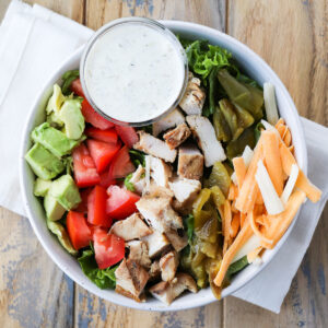 prepared salad in a bowl with grilled chicken, avocado, tomato, hatch green chiles, shredded cheese and a side of spicy ranch