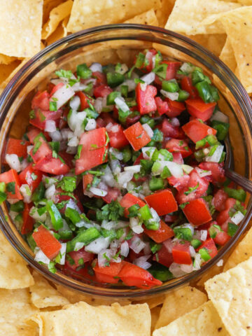 The bowl is filled with a vibrant mix of diced tomatoes, onions, jalapeños, and cilantro. The ingredients are evenly distributed, and the colors are bright and contrasting. The texture is chunky and there are visible pieces of tomato and onion. The cilantro leaves are finely chopped and scattered throughout