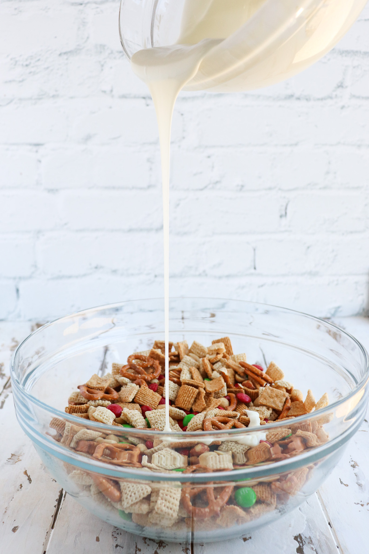 pouring melted premier white chocolate chips into a large glass bowl of cereal, pretzels, peanuts and m&ms