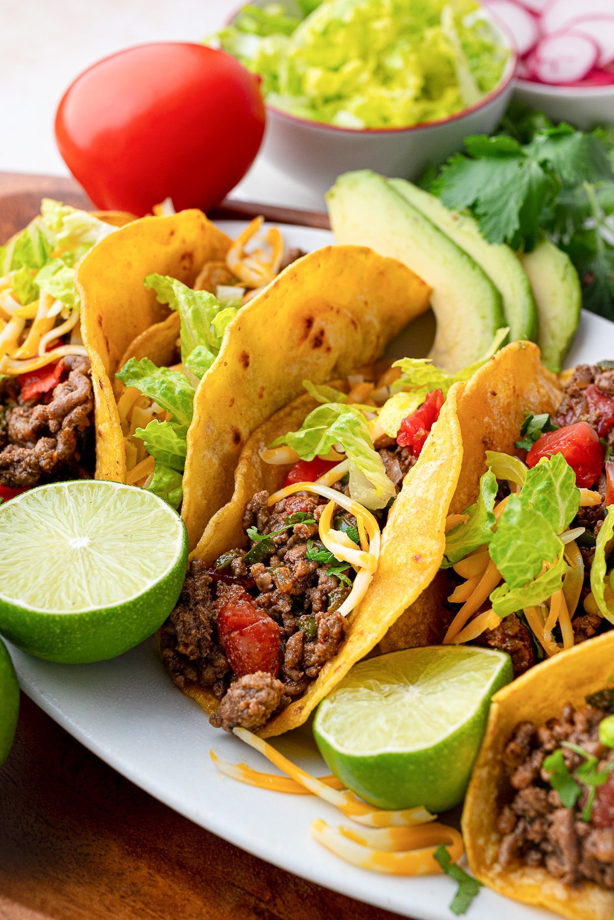 crispy beef taco meat in shells garnished with Roma tomatoes, limes, cilantro and cheese