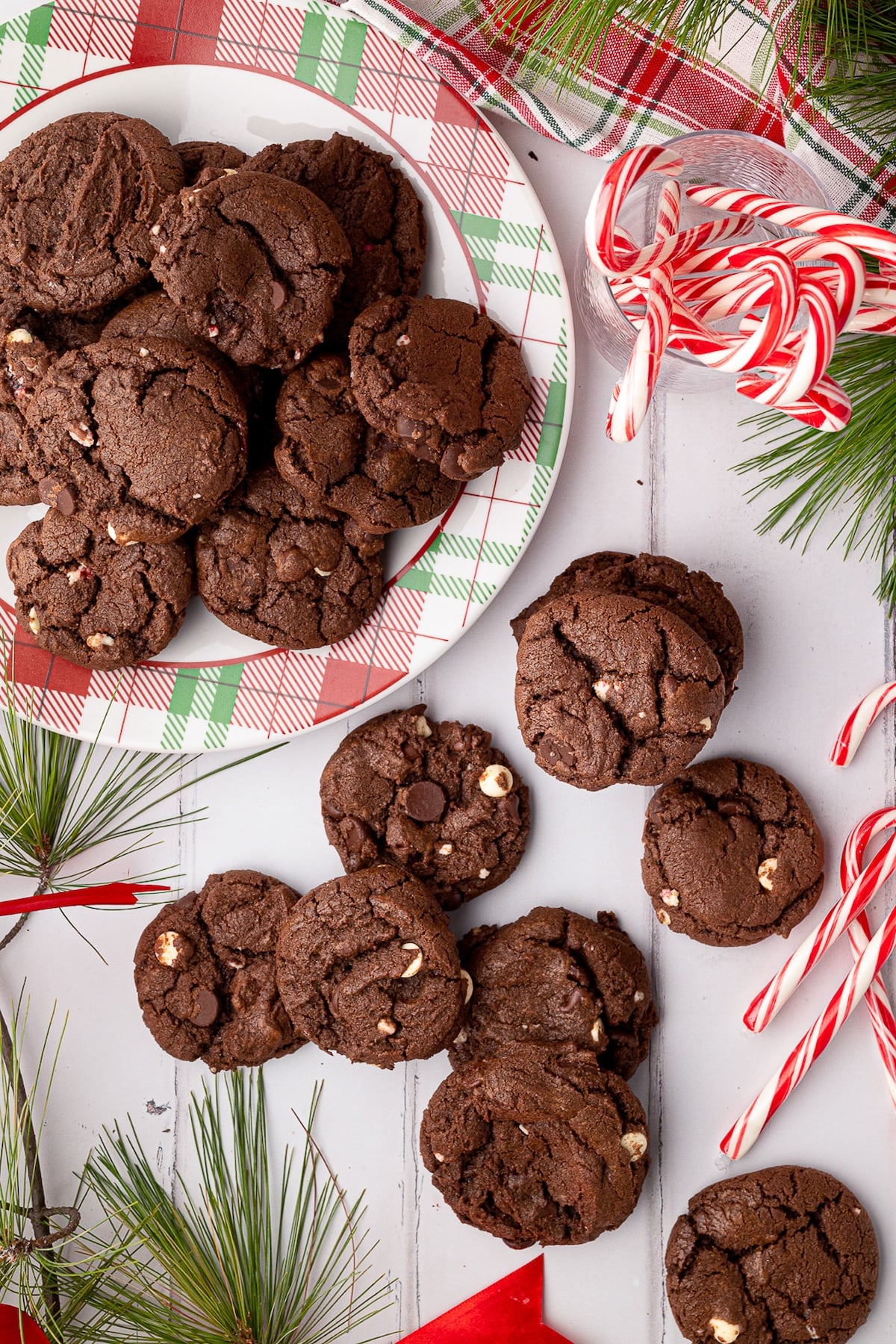 Festive scene of baked Chocolate Peppermint Cookies on a white board, surrounded by plaid towel, greenery and candy canes.