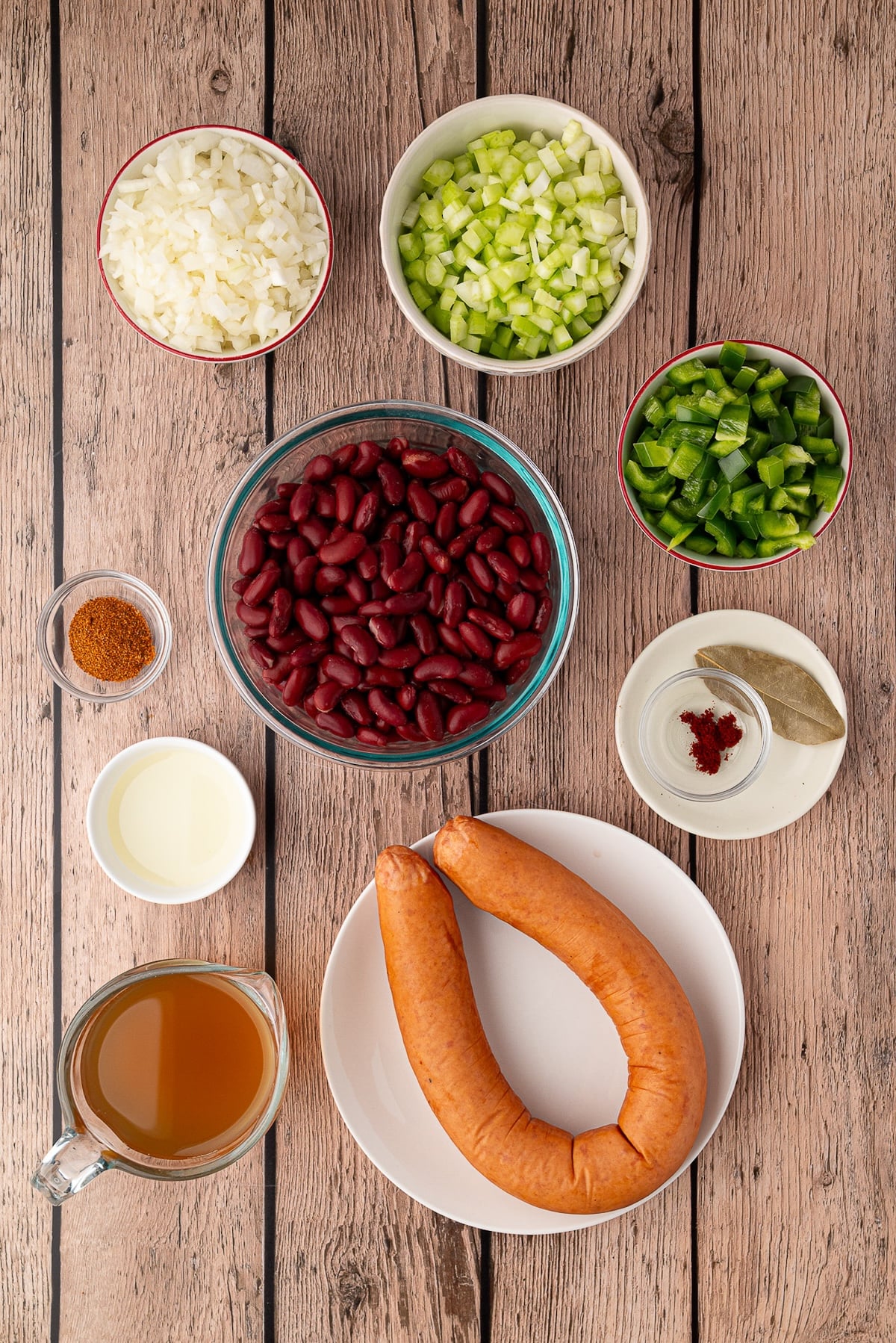 Ingredients for easy skillet red beans including canned kidney beans, H-E-B smoked sausage, diced onion, green bell pepper, celery, and Cajun spices arranged on a table.