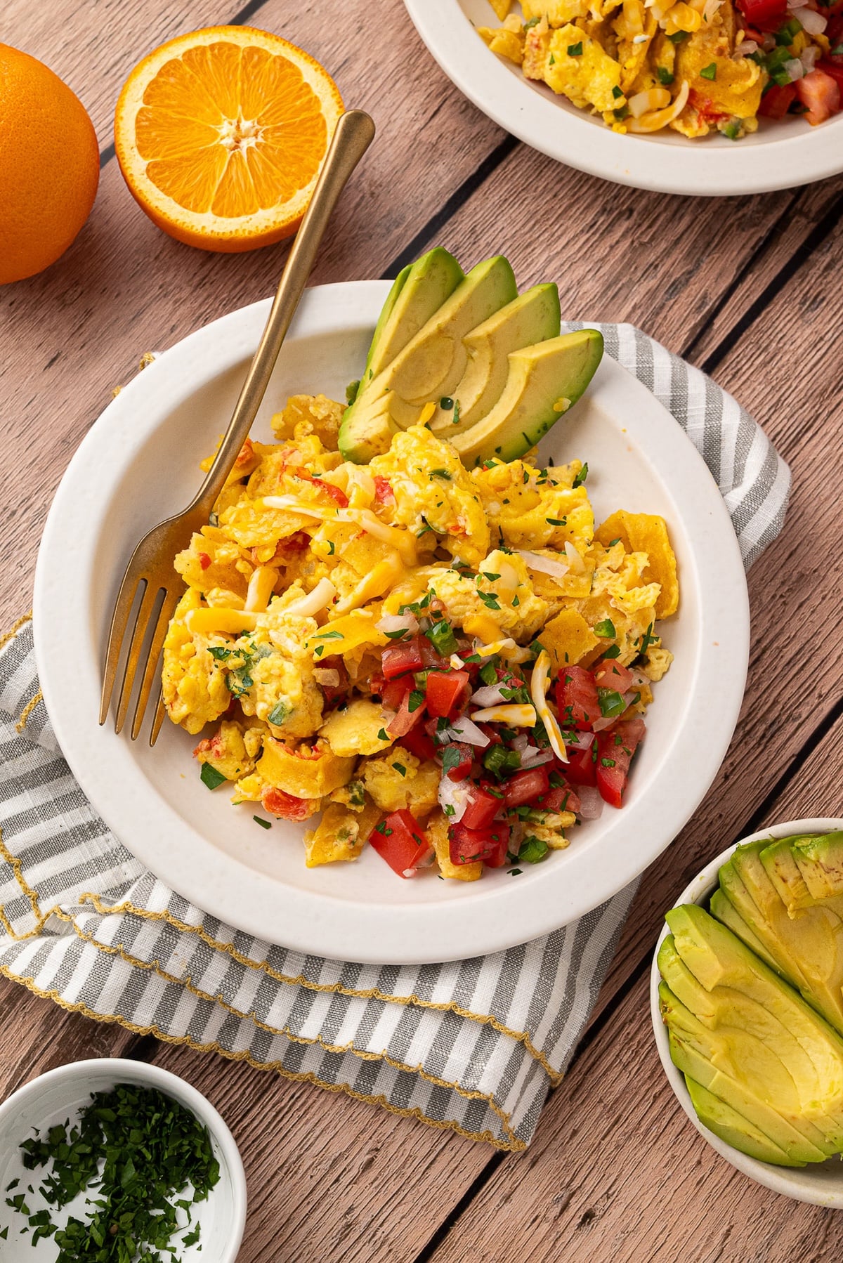 Plated Migas served on a white ceramic dish, showcasing the vibrant colors of yellow scrambled eggs, bright red tomatoes, and deep green sliced avocado. Light reflects off the melted cheese and crispy tortilla strips.