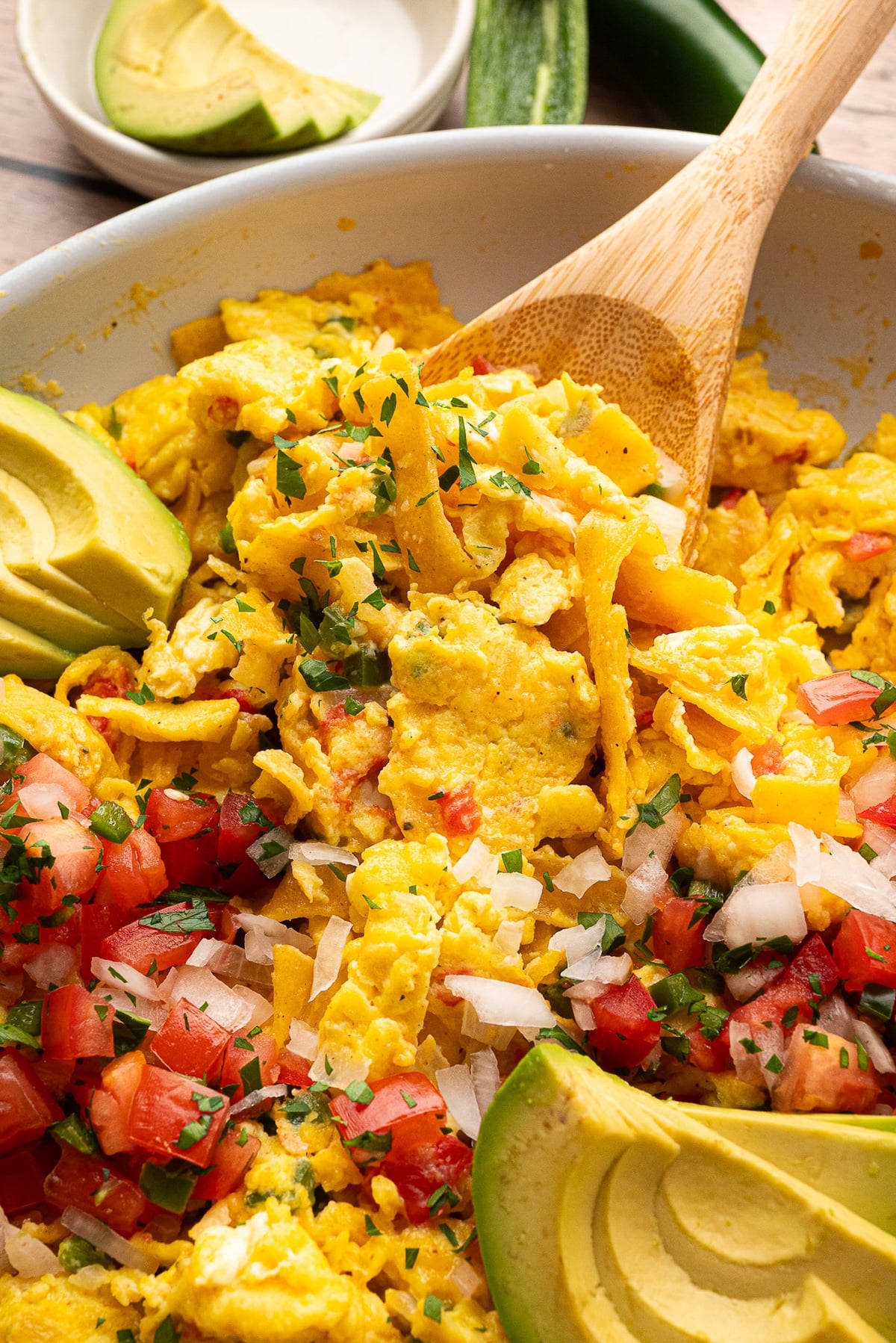 A close-up overhead view of Tex-Mex Migas in a skillet, featuring scrambled eggs mixed with crispy corn tortilla strips, melted cheddar cheese, diced tomatoes, and onions. Garnished with fresh cilantro and sliced avocado.