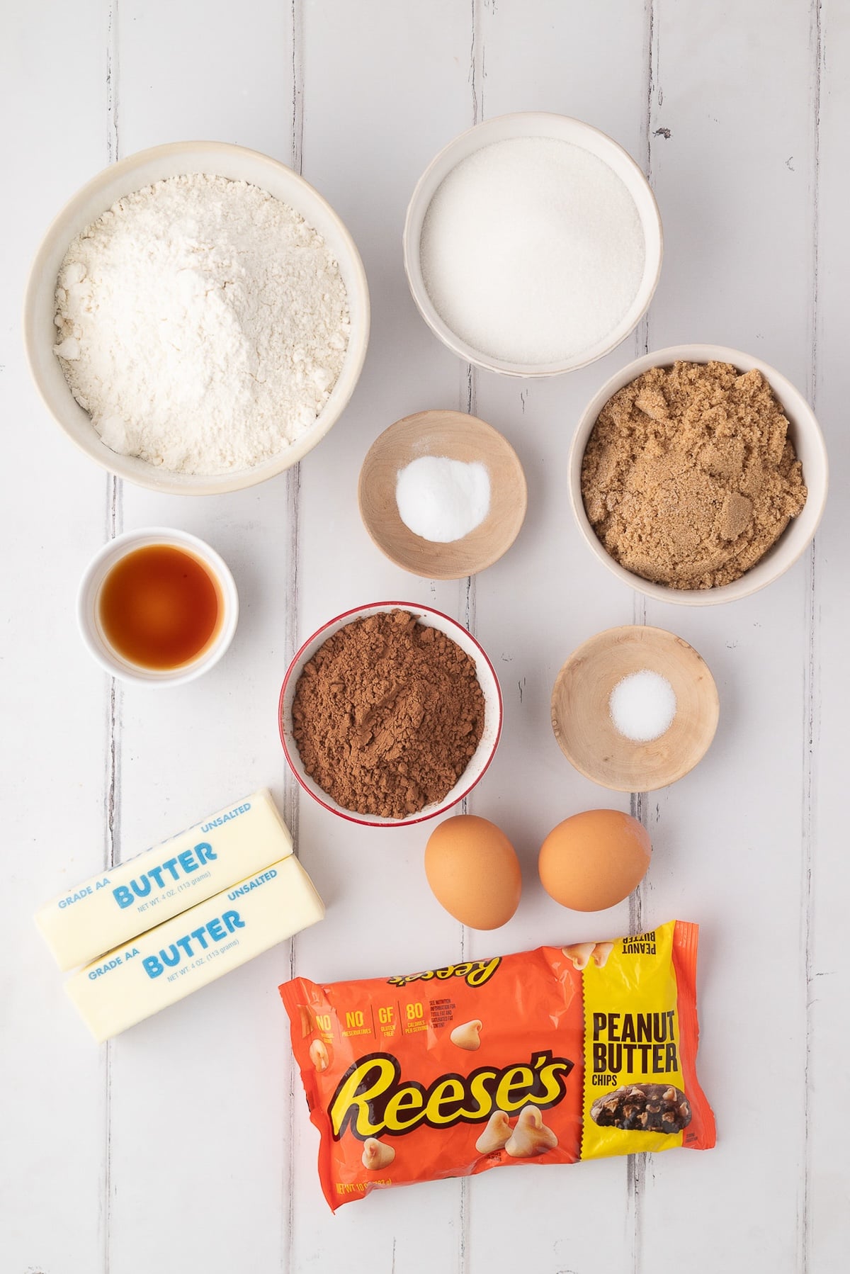 Overhead view of ingredients for chocolate peanut butter chip cookies, including unsweetened cocoa powder, flour, Reese's peanut butter chips, butter, and eggs on a white counter.