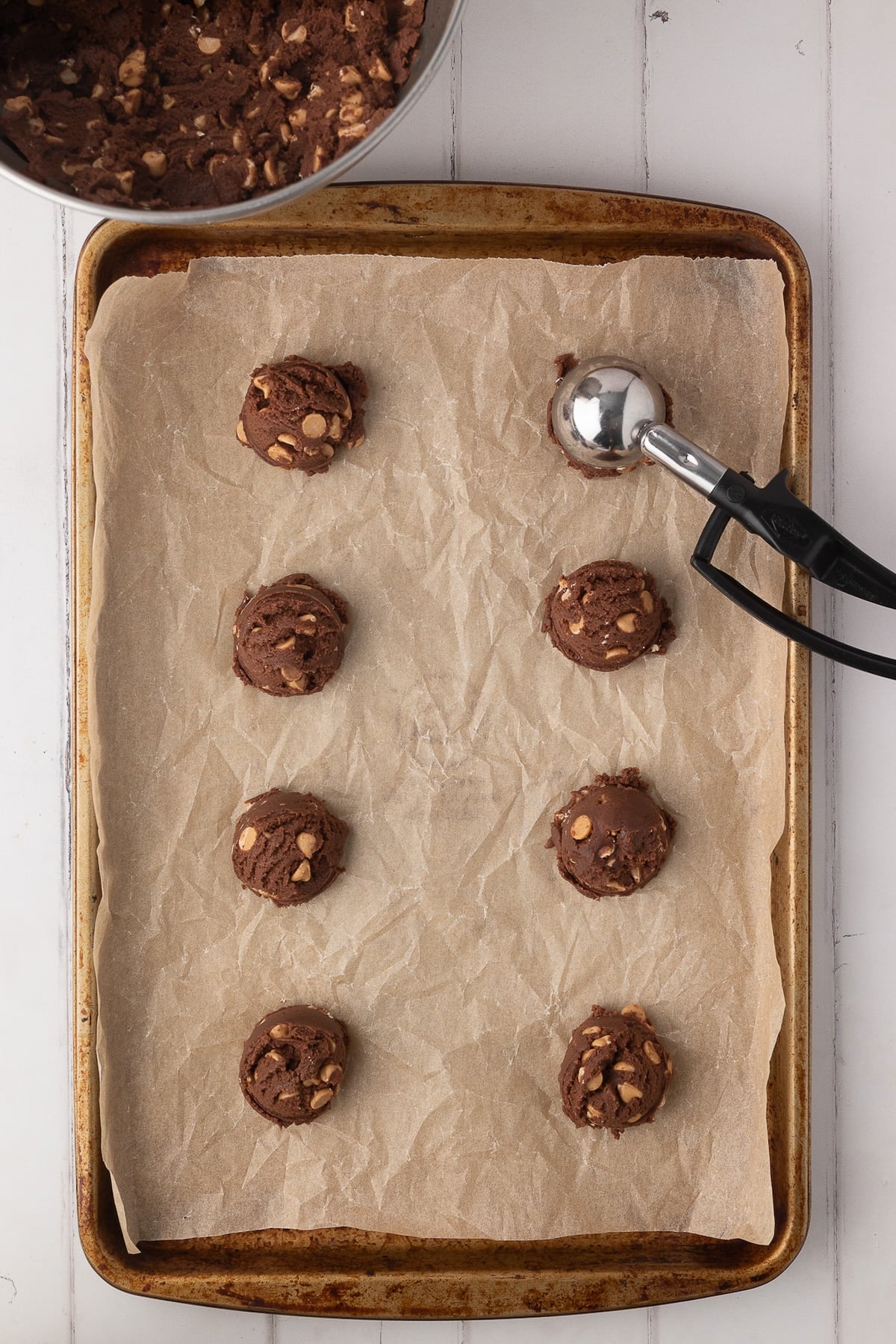 Using a standard-sized metal cookie scoop to portion out chocolate dough onto a parchment-lined baking sheet.