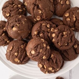 A large white ceramic plate piled high with soft and chewy chocolate peanut butter chip cookies, showing the rich cocoa texture and creamy Reese's peanut butter chips throughout.