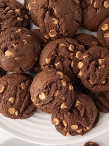A large white ceramic plate piled high with soft and chewy chocolate peanut butter chip cookies, showing the rich cocoa texture and creamy Reese's peanut butter chips throughout.