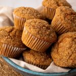 A bowl of freshly baked, healthy whole wheat carrot muffins with crunchy turbinado sugar tops, perfect for a Spring or Easter breakfast.