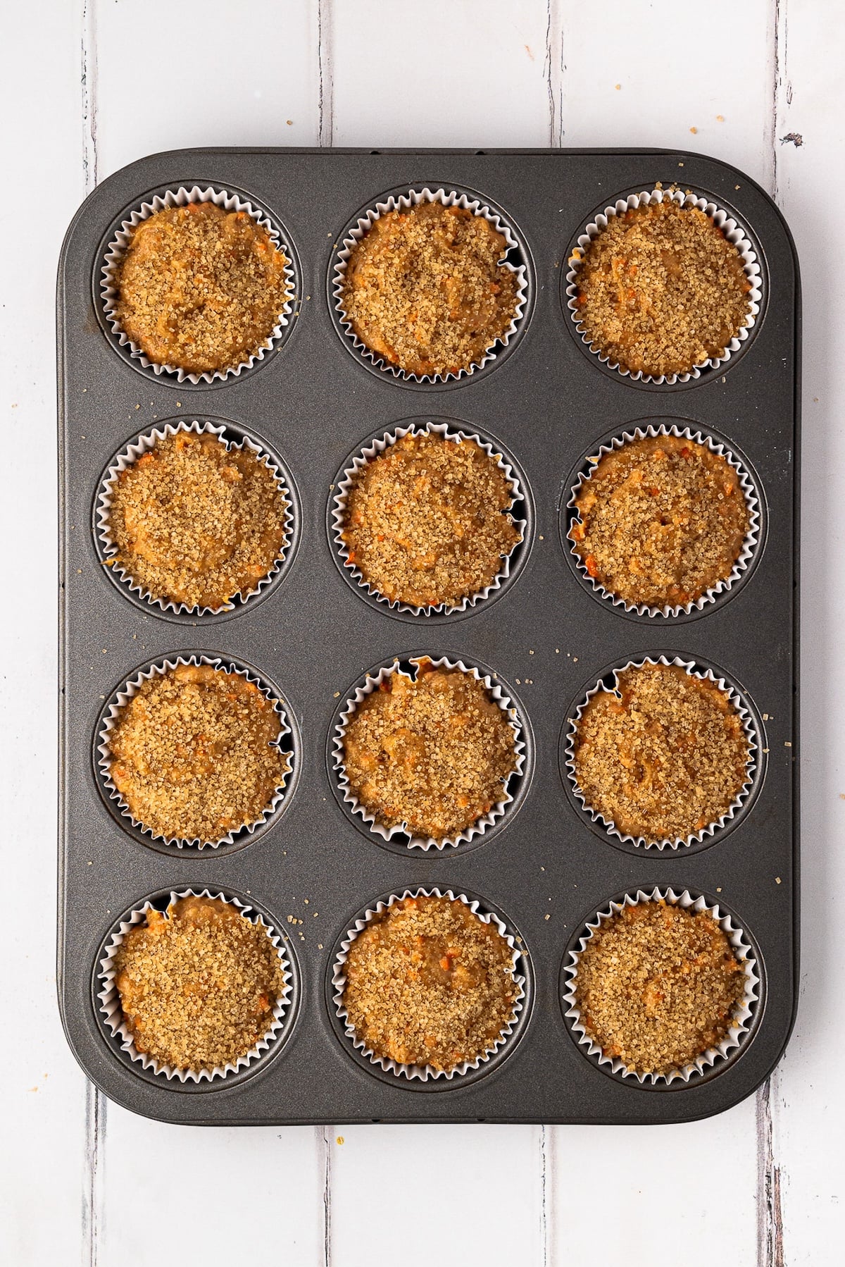 Muffin batter in a muffin tin being sprinkled with coarse turbinado sugar for a bakery-style crunch.