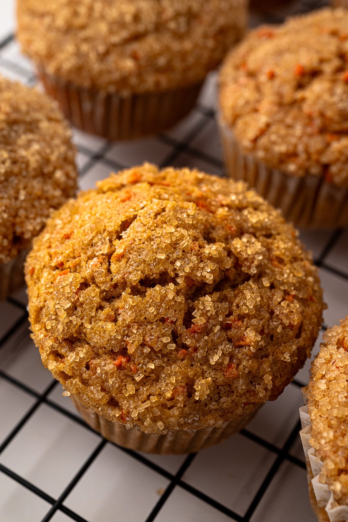 A close-up of a golden-brown whole wheat carrot muffin on a wire rack, showing the crunchy turbinado sugar crust and visible shreds of fresh orange carrots.