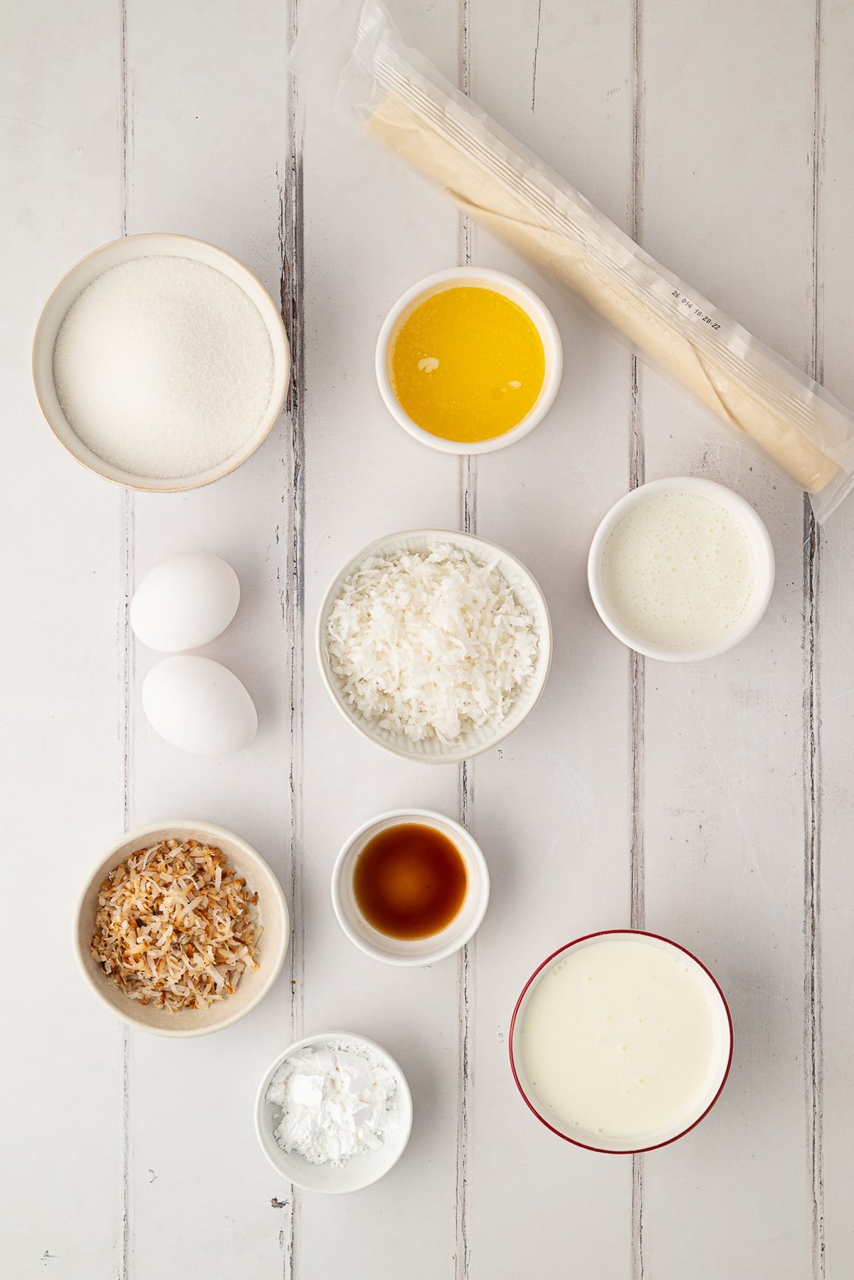 Ingredients needed to make an easy coconut pie, including a bowl of shredded coconut, an unbaked pie crust, buttermilk, sugar, butter, and eggs arranged on a kitchen counter.