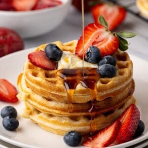 Maple syrup being poured on a stack of thick and fluffy buttermilk waffles, garnished with fresh berries.