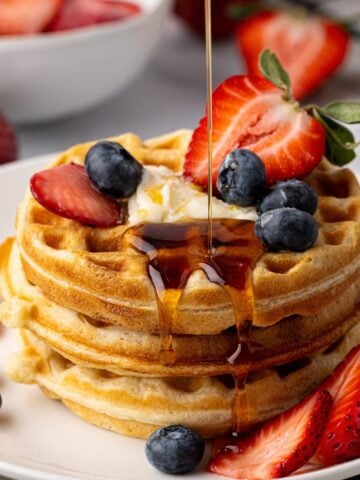 Maple syrup being poured on a stack of thick and fluffy buttermilk waffles, garnished with fresh berries.