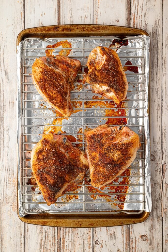 Freshly baked seasoned split chicken breasts resting on a baking sheet with crispy skin and clear pan juices.