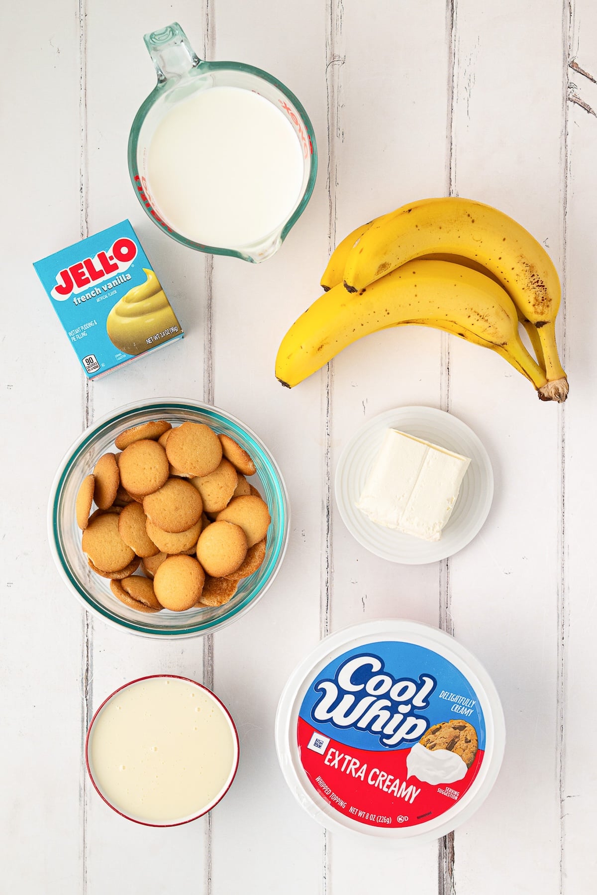 Ingredients for no-bake banana pudding laid out on a counter, including French vanilla pudding mix, 2% milk, sweetened condensed milk, softened cream cheese, fresh bananas, whipped topping, and vanilla wafers.