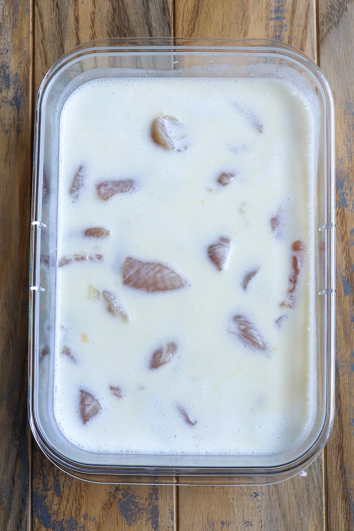 Bite-sized pieces of raw chicken marinating in a bowl of creamy buttermilk and whisked eggs, with bubbles rising to the surface.