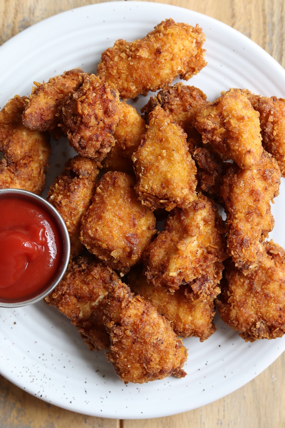 A plate filled with golden-brown, extra-crispy cornflake fried chicken tenders, served on a rustic wooden table with a small bowl of dipping sauce.