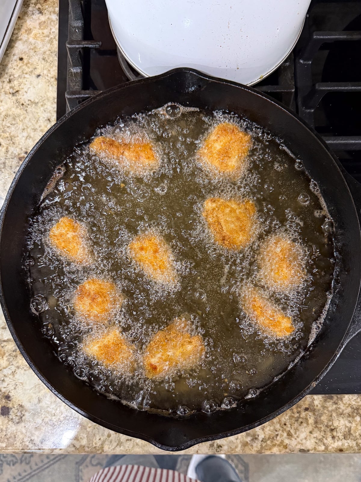 Several pieces of cornflake fried chicken frying in hot, bubbling oil within a deep cast-iron skillet, turning a beautiful golden-brown color.