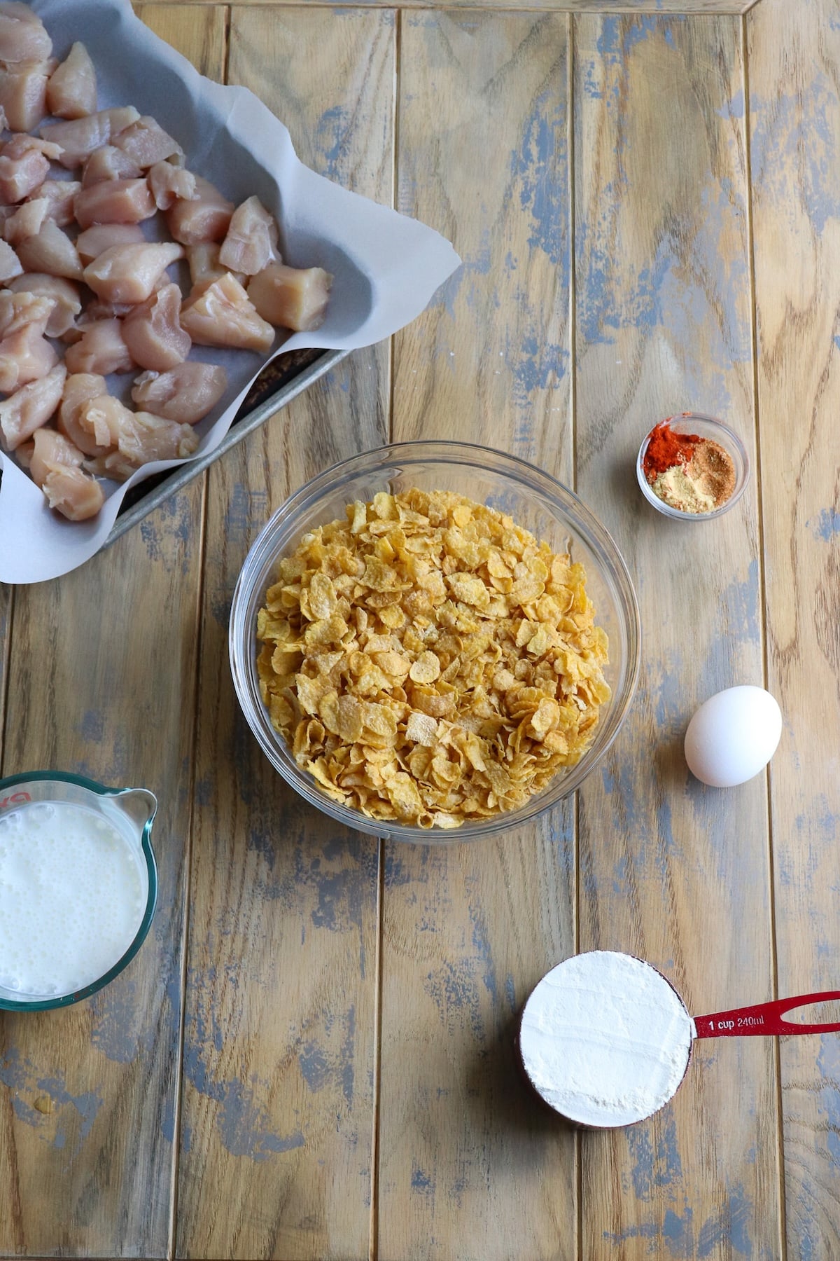 An overhead view of all ingredients for the cornflake fried chicken, including raw chicken pieces, a bowl of buttermilk, eggs, a bag of crushed cornflakes, flour, and seasonings on a kitchen counter.