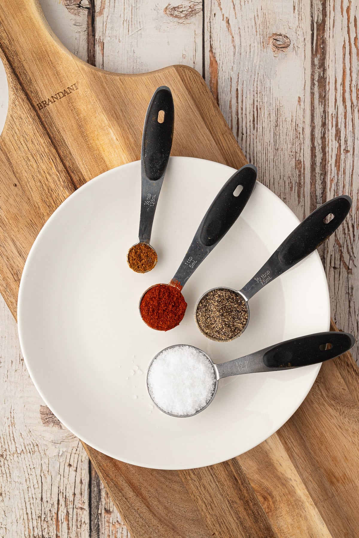 Individual spoons of spices including paprika, garlic powder, salt, and black pepper laid out to make a savory Texas dry rub for chicken.
