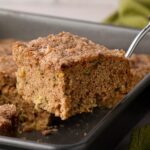 A metal spatula lifting a square slice of moist cinnamon zucchini coffee cake out of a baking pan, revealing a soft, plush buttermilk crumb and cinnamon sugar topping.
