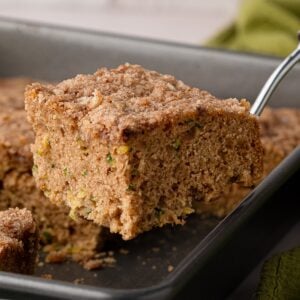 A metal spatula lifting a square slice of moist cinnamon zucchini coffee cake out of a baking pan, revealing a soft, plush buttermilk crumb and cinnamon sugar topping.