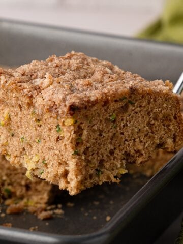A metal spatula lifting a square slice of moist cinnamon zucchini coffee cake out of a baking pan, revealing a soft, plush buttermilk crumb and cinnamon sugar topping.