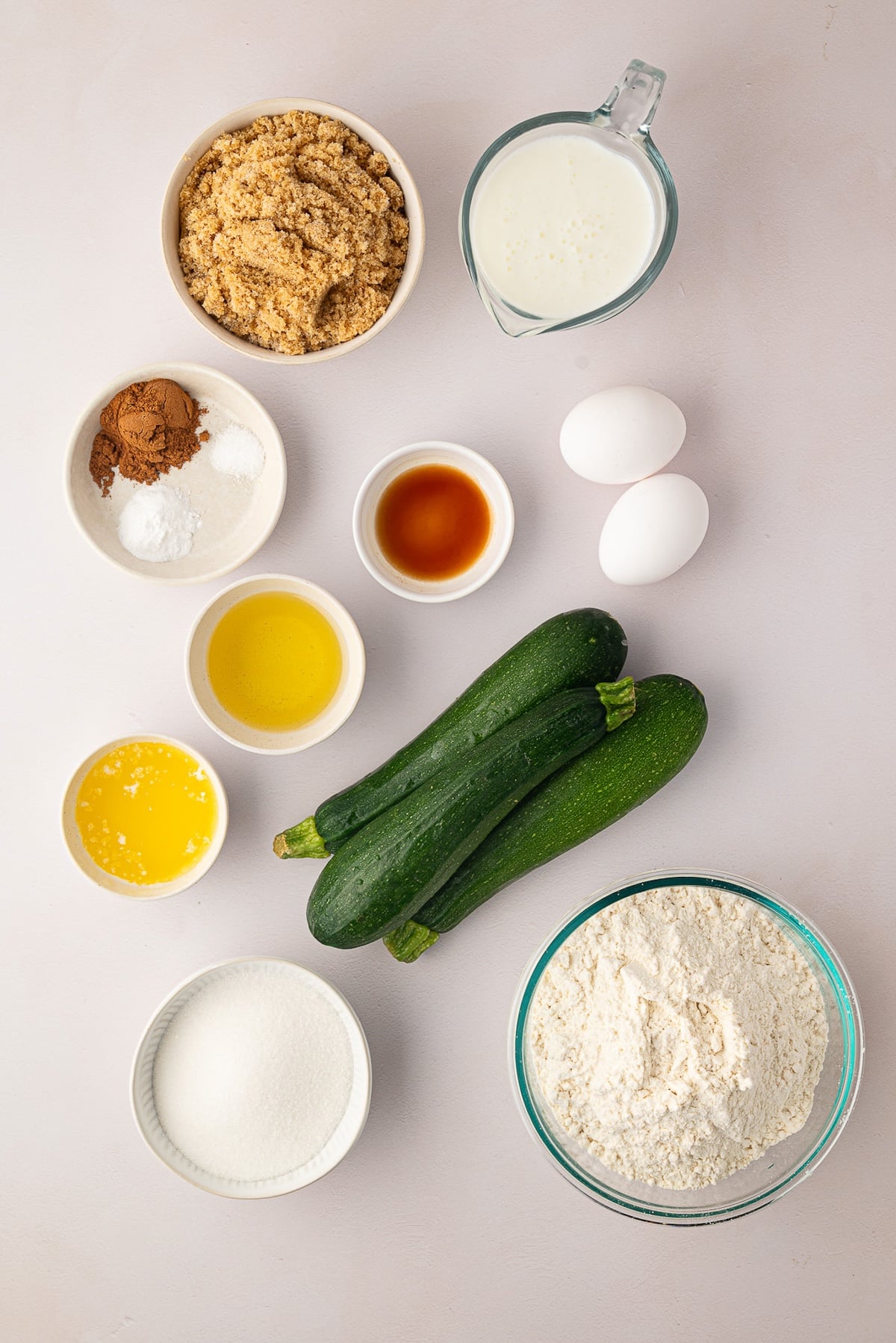 An overhead shot of ingredients for cinnamon zucchini coffee cake spread out on a kitchen counter, including brown sugar, grated zucchini, buttermilk, flour, eggs, and cinnamon.