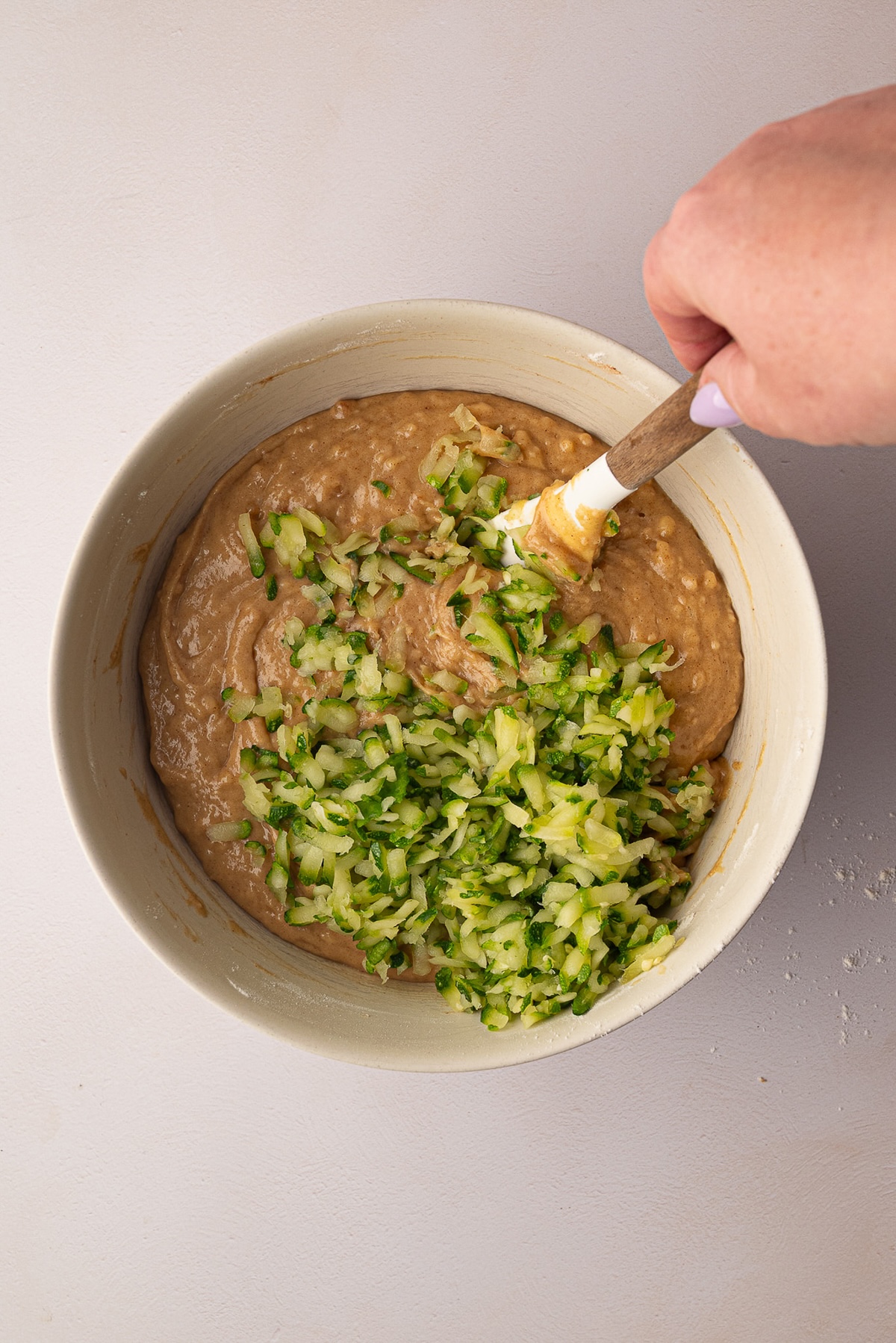 A silicone spatula folding freshly grated green zucchini shreds into a thick, creamy cake batter in a mixing bowl.