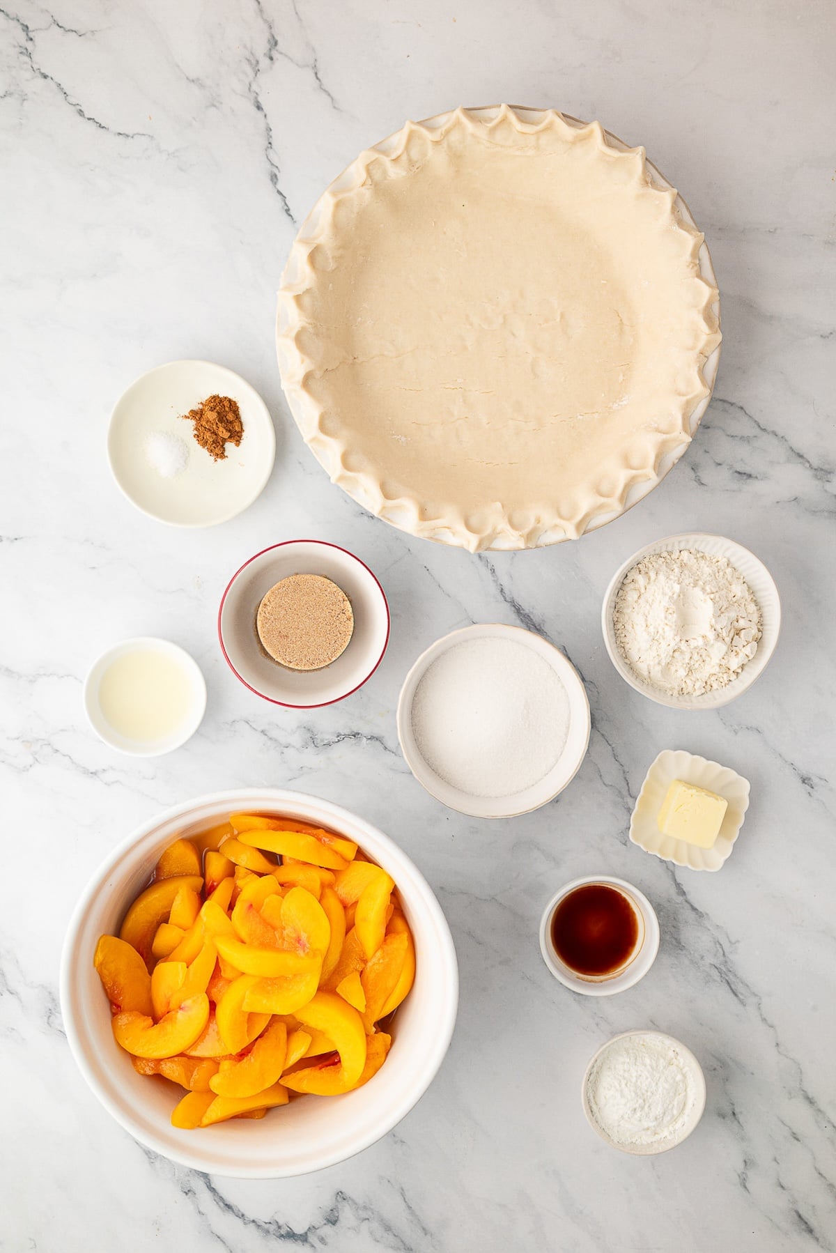 An overhead shot of the ingredients for a homemade peach crumble pie, including sliced peaches, light brown sugar, cinnamon, flour, butter and a deep dish pie crust.