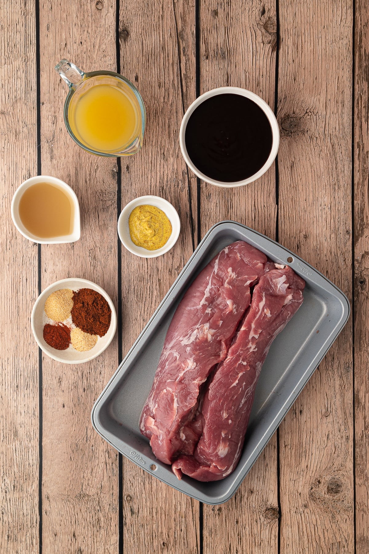 Ingredients for pineapple barbecue pulled pork arranged on a kitchen counter, including raw pork tenderloin, a can of pineapple juice, chicken broth, BBQ sauce, yellow mustard, and dried spices.