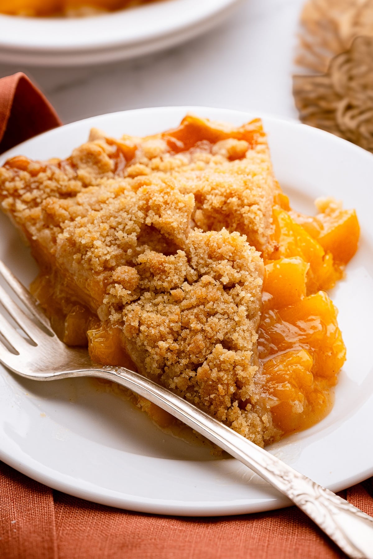 A clean slice of the best peach crumble pie on a small white dessert plate, showing the thick layers of velvety peach filling and crisp topping.