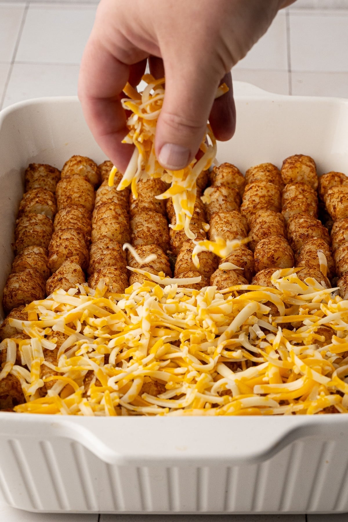 Sprinkling freshly shredded cheddar cheese evenly over crispy, golden-baked tater tots in a casserole dish.