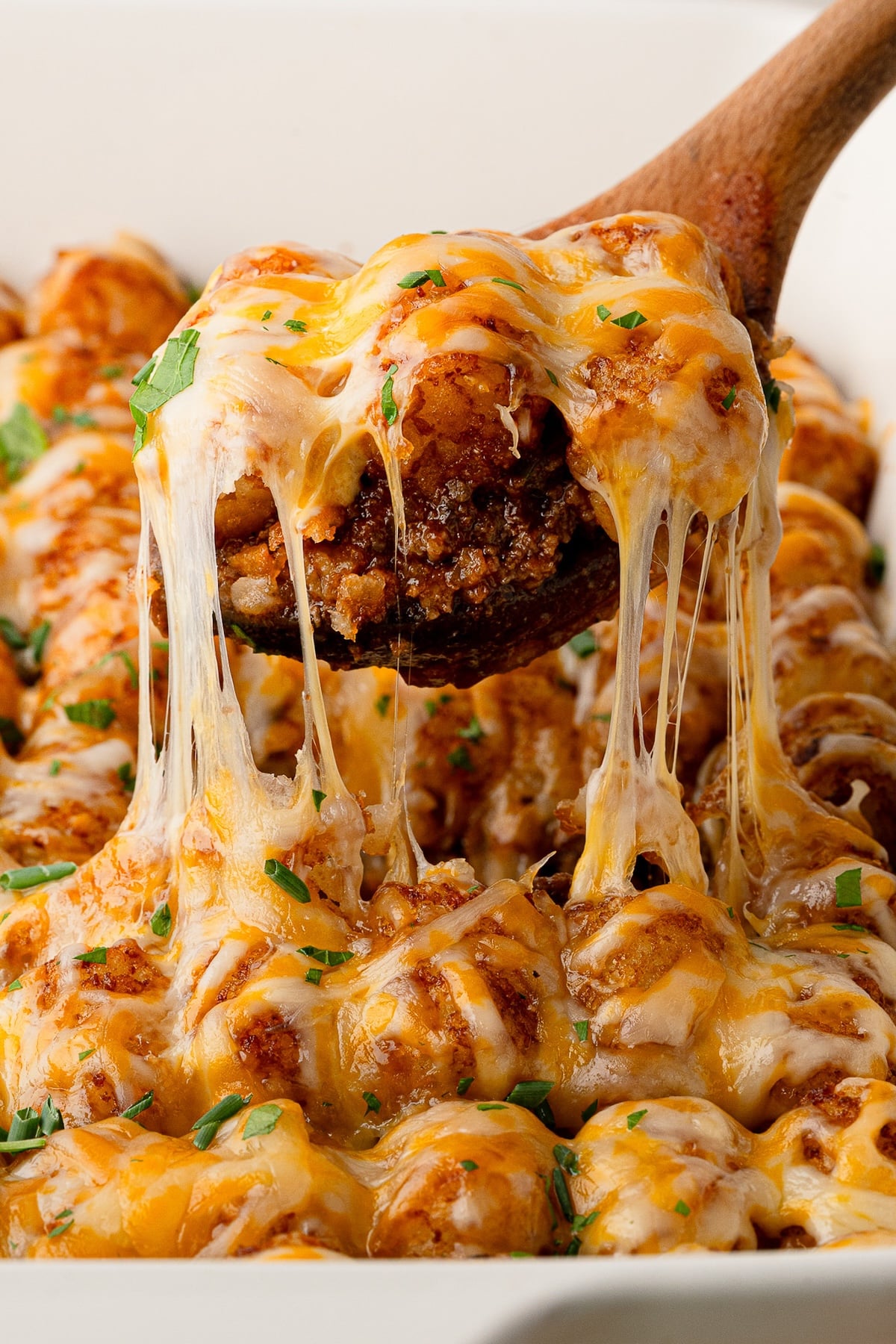 Wooden spoon lifting a serving of Sloppy Joe Tater Tot Casserole out of a baking dish, showing a stretchy cheese pull, crispy tots, and rich ground beef base.