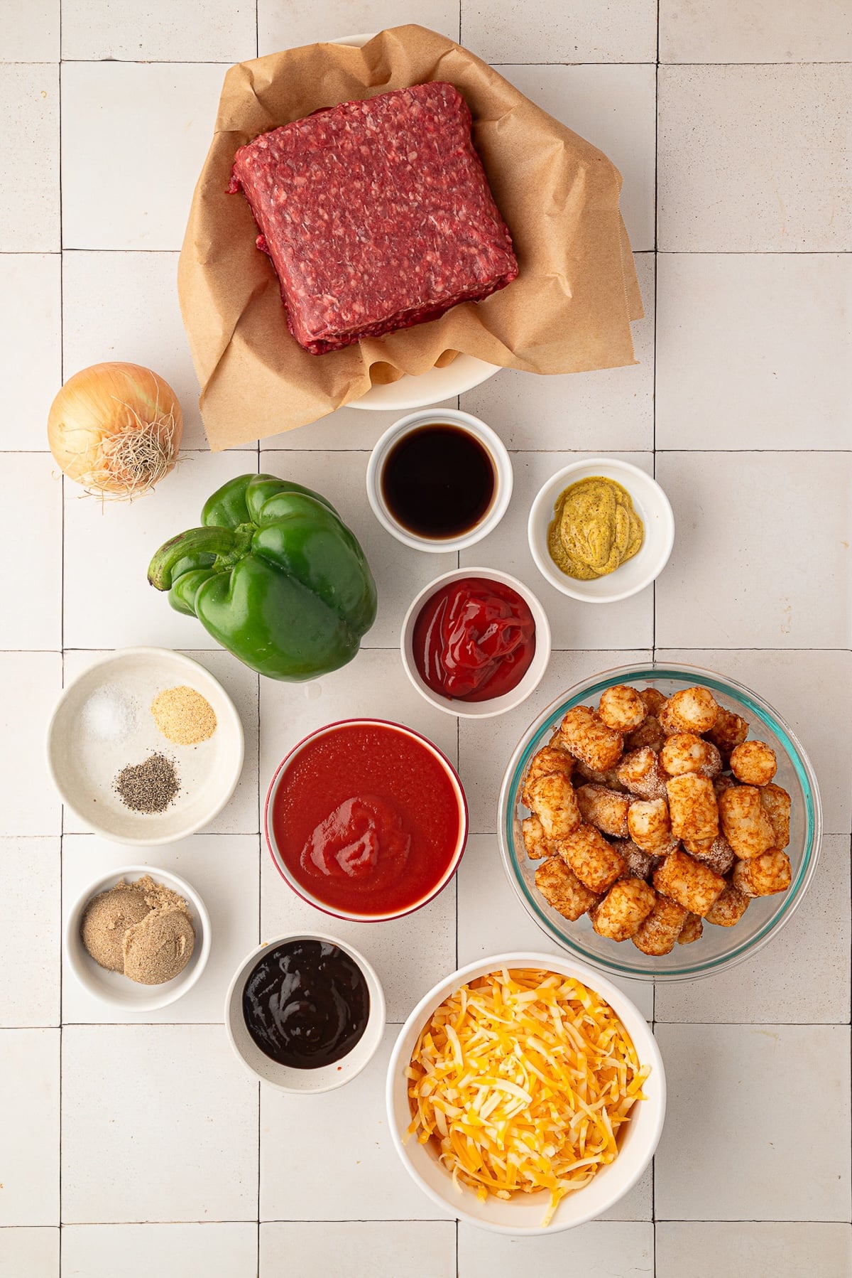 Ingredients for Sloppy Joe Tater Tot Casserole arranged on a counter, including ground beef, diced green bell pepper, chopped onion, canned tomato sauce, BBQ sauce, ketchup, spices, brown sugar, frozen tater tots, and shredded cheddar cheese.