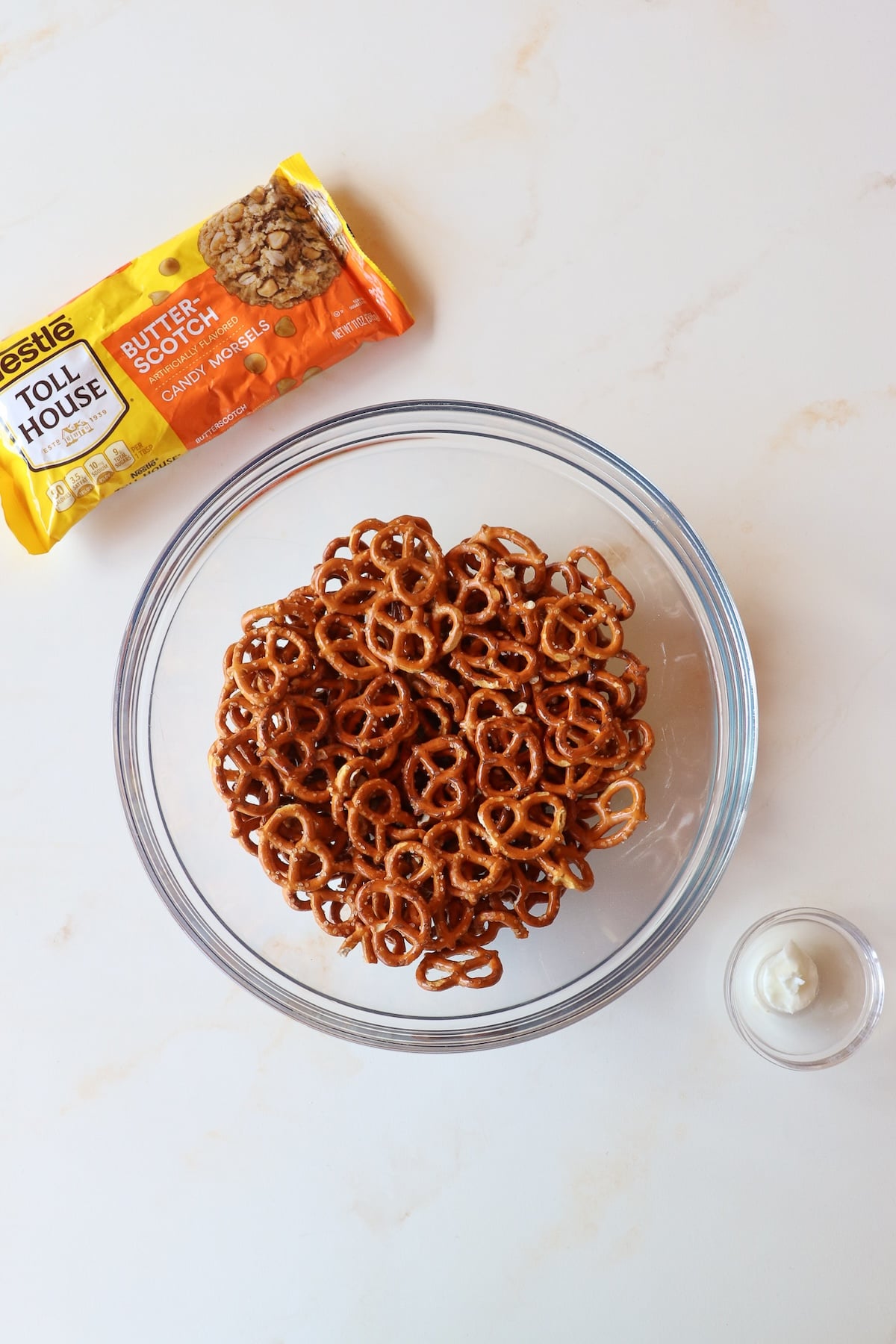 A glass bowl of mini twist pretzels, butterscotch chips, and shortening on a countertop.