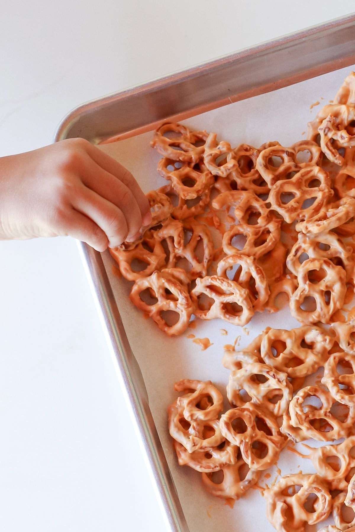 A child's hand takes a butterscotch coated pretzel from a parchment lined sheet pan.